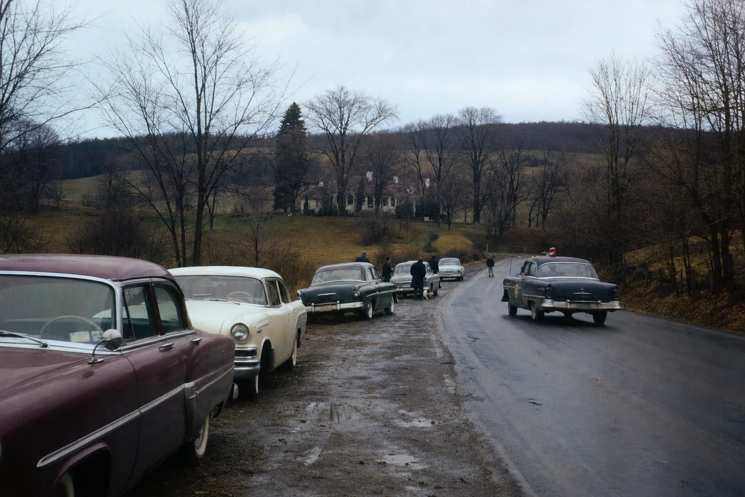 Photorealistic editorial reconstruction of a rural 1957 road with period cars, police, and a distant farmhouse